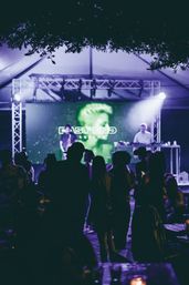 Silhouetted crowd at an outdoor nighttime music event under a tent, watching a DJ and vocalist on a purple-green lit stage with a large LED backdrop.