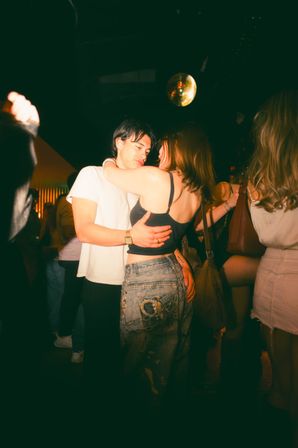 Couple embracing and dancing under a disco ball on a dimly lit nightclub dance floor, surrounded by other partygoers.