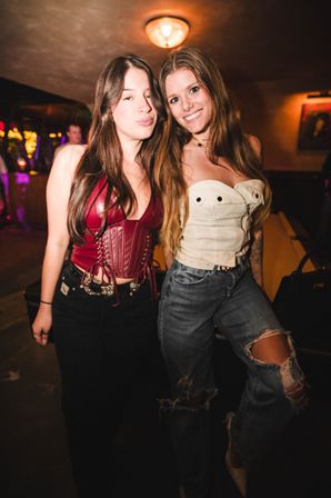 Two women posing in a dimly lit bar or nightclub — one in a red leather corset and black jeans, the other in a white corset and ripped jeans — smiling under warm overhead light with colorful string lights in the background.