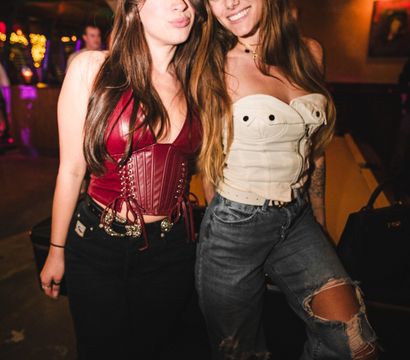 Two women posing in a dimly lit bar or nightclub — one in a red leather corset and black jeans, the other in a white corset and ripped jeans — smiling under warm overhead light with colorful string lights in the background.