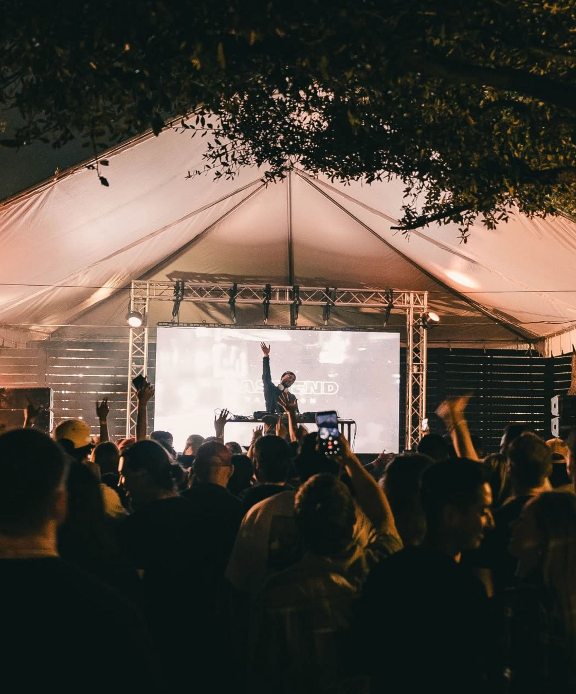 Outdoor night concert under a large white tent with a DJ on stage raising a hand, bright screen and stage lights, lively crowd with phones raised beneath tree branches.