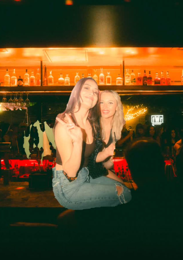 Two friends perched on a bar counter smiling and flashing a peace sign in a lively city nightclub, illuminated bottles on the back shelf and a crowded, warmly lit dance floor.