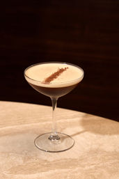 Espresso martini cocktail in a coupe glass with creamy foam and a diagonal cocoa dust garnish, set on a marble bar table against a dark wood background
