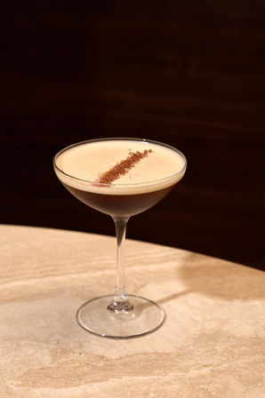 Espresso martini cocktail in a coupe glass with creamy foam and a diagonal cocoa dust garnish, set on a marble bar table against a dark wood background