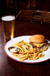 Pub-style cheeseburger with melted cheddar and shredded lettuce, piled next to golden herb-sprinkled French fries and a small mayo dip on a white plate, with a pint of beer on a dark wooden table.