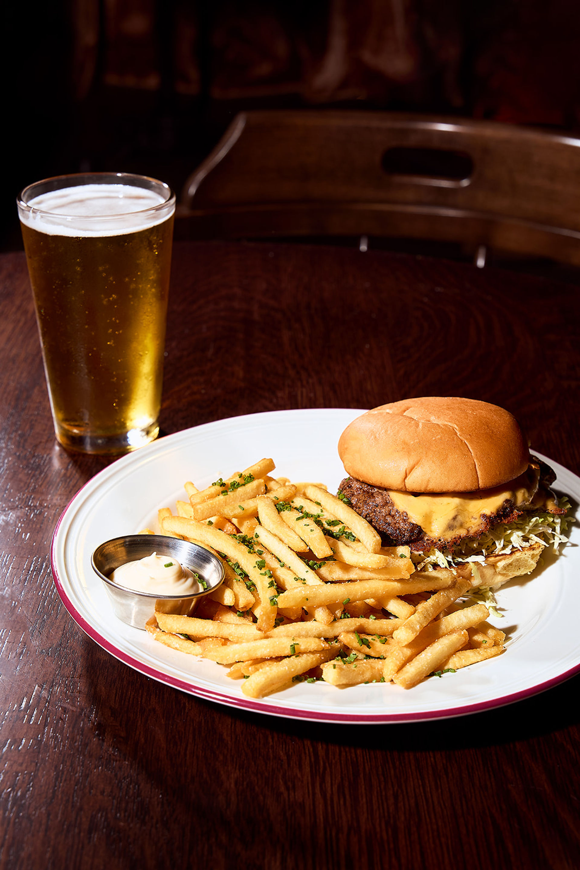 Pub-style cheeseburger with melted cheddar and shredded lettuce, piled next to golden herb-sprinkled French fries and a small mayo dip on a white plate, with a pint of beer on a dark wooden table.