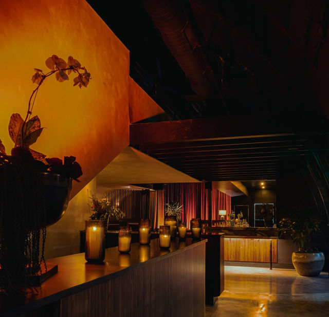 Amber-lit lounge interior with ribbed candle lanterns on a wooden counter, a potted orchid silhouetted against a warm textured wall, velvet drapes and a wood-paneled bar for a cozy urban lounge vibe.