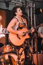 Singer-guitarist on a live concert stage, strumming an acoustic guitar and singing into a mic while wearing paint-splattered overalls and a white tank top, with drums and stage lighting behind him.