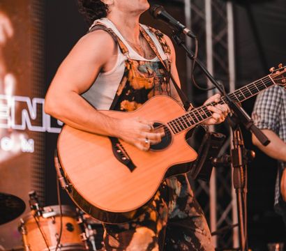 Singer-guitarist on a live concert stage, strumming an acoustic guitar and singing into a mic while wearing paint-splattered overalls and a white tank top, with drums and stage lighting behind him.