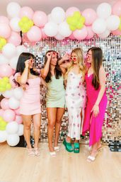 Four women in colorful summer dresses wearing heart-shaped sunglasses pose under a pink, white, and lime balloon arch in front of a mirrored sequin photo-wall backdrop at a festive party.