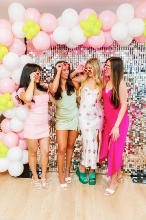Four women in colorful summer dresses wearing heart-shaped sunglasses pose under a pink, white, and lime balloon arch in front of a mirrored sequin photo-wall backdrop at a festive party.