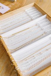 Close-up of delicate gold, silver, and rose-gold chain necklaces neatly arranged in a white foam jewelry display tray on a wooden table, minimalist retail jewelry display.