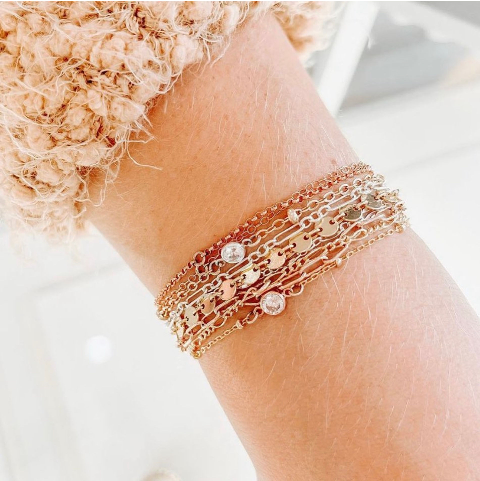 Layered rose gold chain bracelet with small disc charms and bezel-set crystals on a wrist beside a fuzzy cream sweater cuff