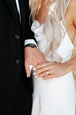 Close-up wedding detail of bride and groom holding hands with wedding rings, bride in white satin dress and long blonde hair, groom in black suit and watch