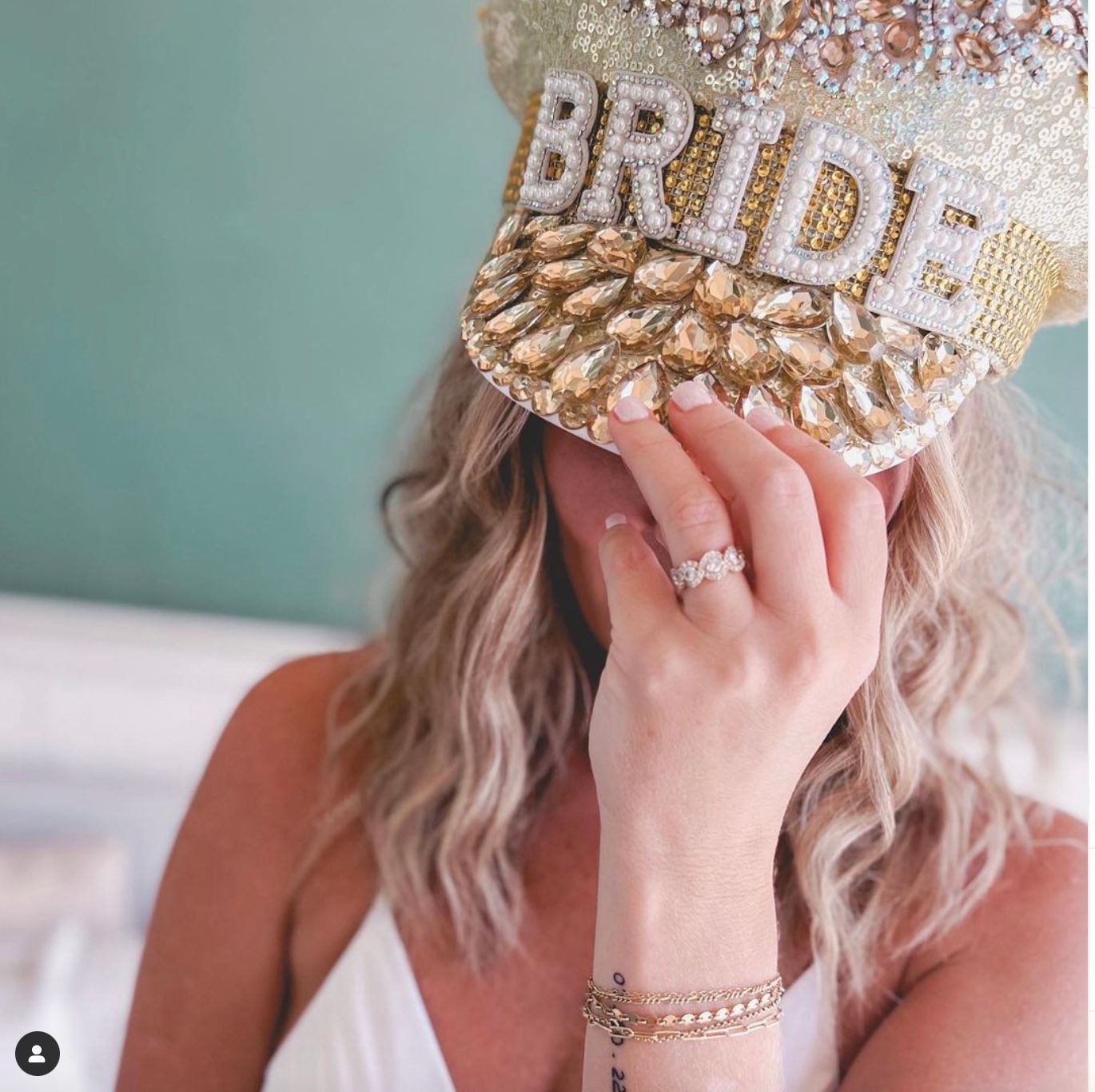 Bride-to-be wearing a gold jeweled cap reading BRIDE, showcasing a sparkling engagement ring, French manicure, layered gold bracelets and wavy blonde hair against a mint background