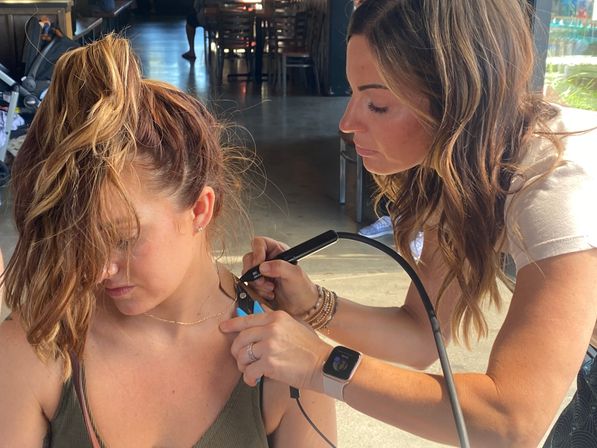 Technician carefully placing small body jewelry near a woman’s collarbone while the client with a messy ponytail looks down in a sunlit cafe-style space, technician using pliers and an electronic tool.