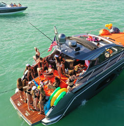 Aerial view of a summer yacht party in turquoise water — a crowded luxury boat with people in bikinis on orange deck cushions, inflatables, balloons, food and an American flag.