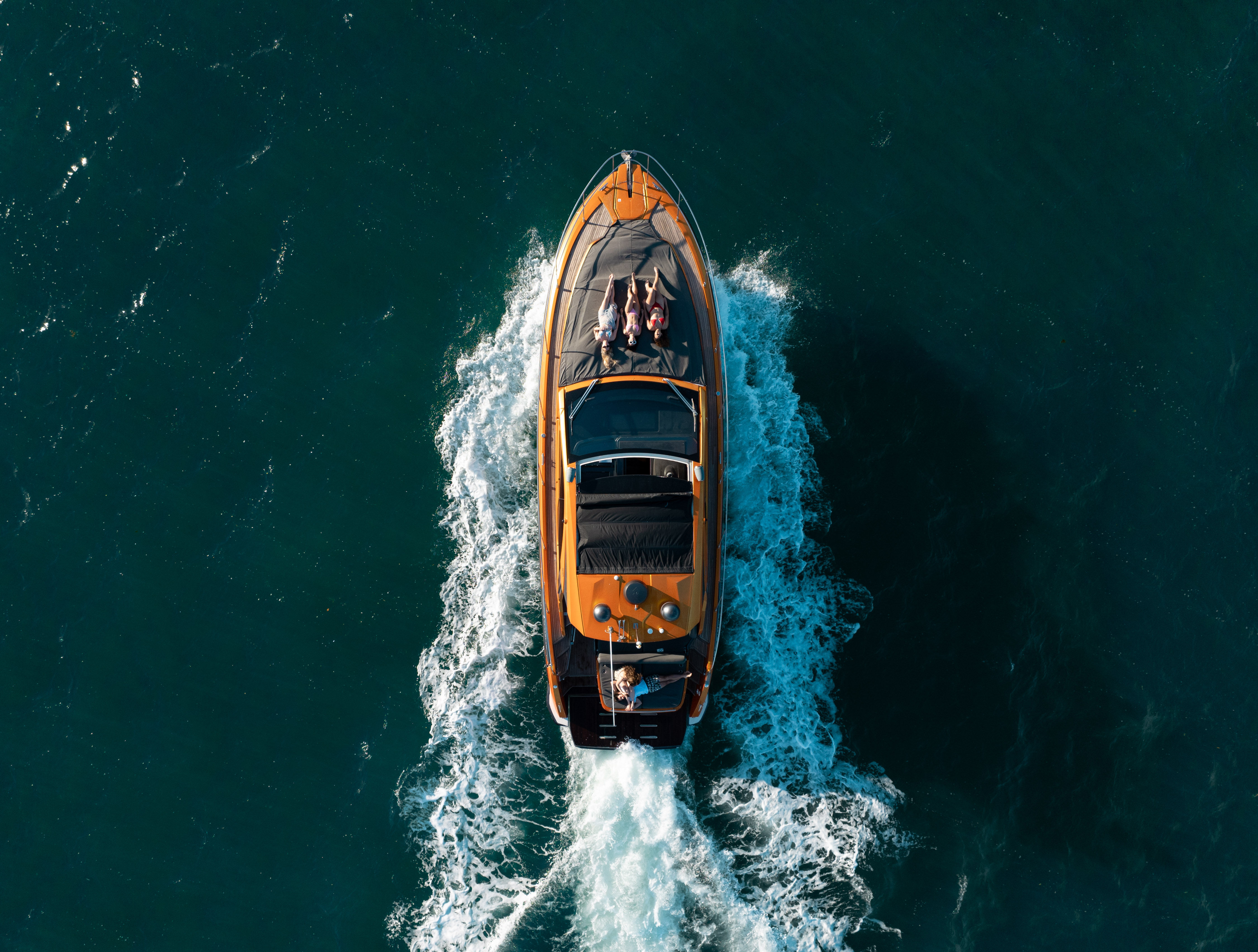 Aerial drone view of an orange luxury motorboat cruising through deep blue ocean with a white wake behind; two people lounging on the foredeck and a driver at the helm.