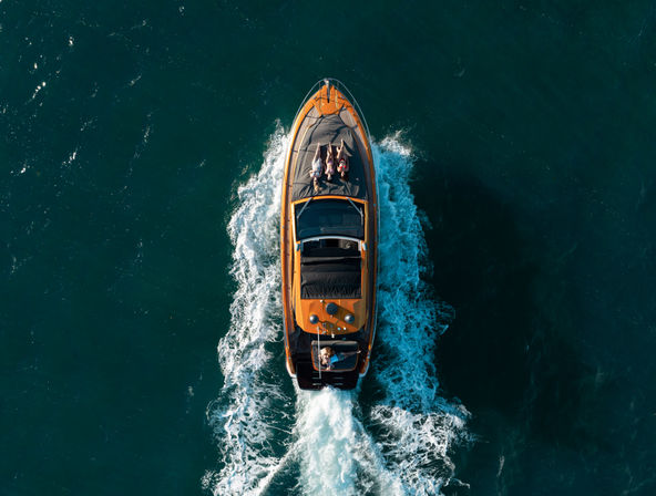 Aerial drone view of an orange luxury motorboat cruising through deep blue ocean with a white wake behind; two people lounging on the foredeck and a driver at the helm.