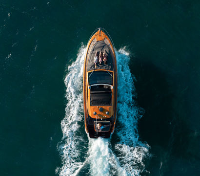 Aerial drone view of an orange luxury motorboat cruising through deep blue ocean with a white wake behind; two people lounging on the foredeck and a driver at the helm.