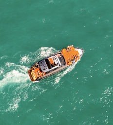 Aerial view of a luxury yacht cutting through turquoise water, passengers sunbathing on bright orange deck cushions amid colorful balloons.