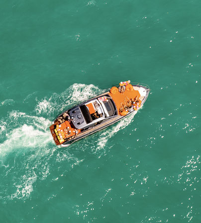Aerial view of a luxury yacht cutting through turquoise water, passengers sunbathing on bright orange deck cushions amid colorful balloons.