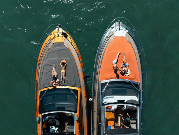 Aerial view of two motorboats tied side-by-side in emerald water, with sunbathers relaxing on bright orange and gray foredecks—summer boating scene with people in swimsuits soaking up the sun.