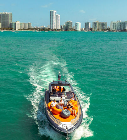 Motorboat cruising turquoise water toward a beachfront city skyline on a sunny day, passengers lounging on bright orange beanbag seats at the bow
