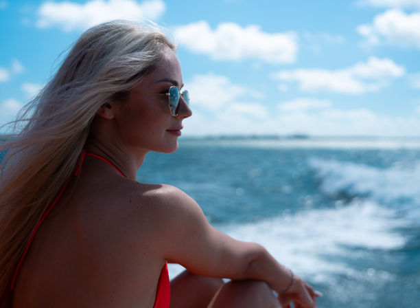 Blonde woman in sunglasses and red bikini gazing toward the horizon from a boat over a sparkling blue ocean on a sunny day