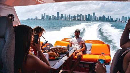 Friends on an orange lounge speedboat cruising away from the Miami skyline, table of sandwiches and snacks, a woman snapping a photo and a man relaxing as the boat’s foamy wake stretches across blue water