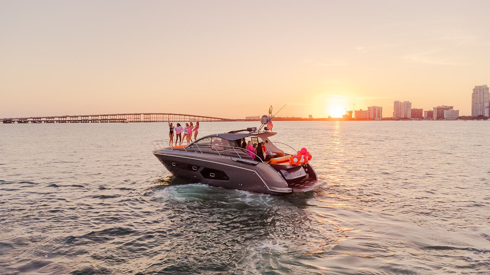 Luxury motor yacht with friends and pink balloons cruising on calm water toward a coastal city skyline and bridge at sunset.