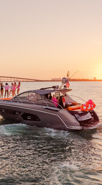 Luxury motor yacht with friends and pink balloons cruising on calm water toward a coastal city skyline and bridge at sunset.