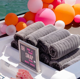 Rolled gray towels on a boat deck beside bright pink and orange balloons and a playful letterboard sign — festive nautical party setup