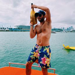 Shirtless man in floral swim trunks and cap holding a golden champagne bottle on an orange boat deck with a cruise ship and Miami skyline in the background.