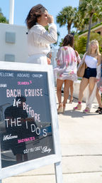Sunny Miami dock with palm trees, women in summer outfits boarding a bachelorette bach cruise next to a chalkboard sign reading "Bach Cruise: Life Before the I Do's".