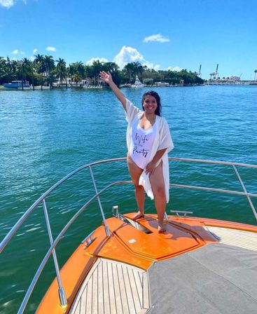 Smiling woman standing on the bow of an orange yacht in turquoise tropical waters, waving with palm-tree lined shoreline and blue sky in the background, wearing a swimsuit that reads 'Wife of the Party'.