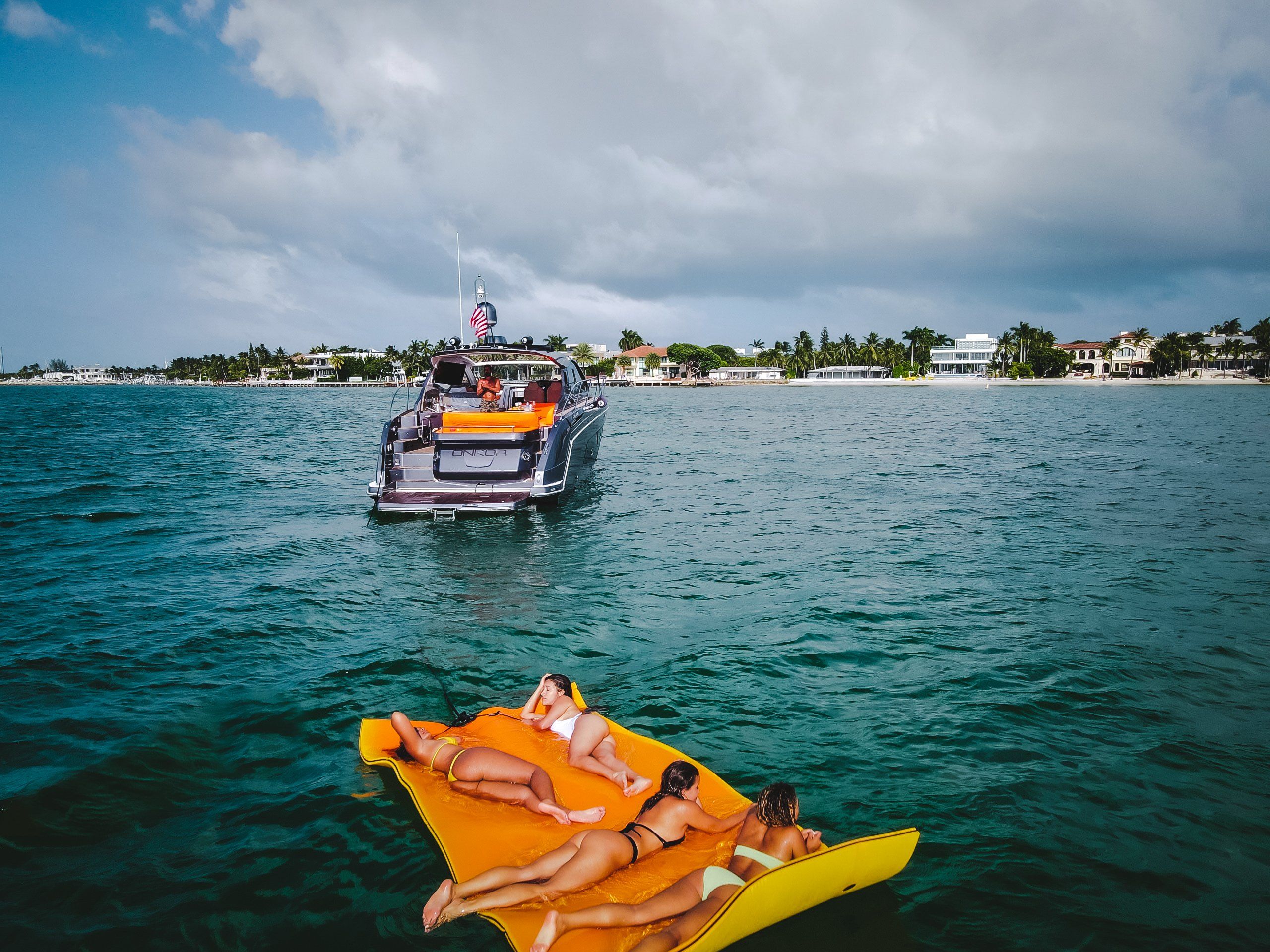 Four people lounging on a bright yellow inflatable mat in turquoise ocean near a luxury yacht off a palm-lined tropical coastline under a cloudy sky