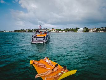 Four people lounging on a bright yellow inflatable mat in turquoise ocean near a luxury yacht off a palm-lined tropical coastline under a cloudy sky