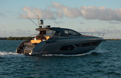 Sleek gray luxury motor yacht cruising on calm coastal waters at golden hour, passengers relaxing on orange aft seating with low shoreline and scattered clouds in the background.