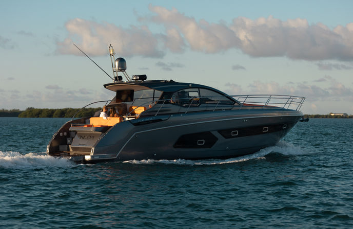 Sleek gray luxury motor yacht cruising on calm coastal waters at golden hour, passengers relaxing on orange aft seating with low shoreline and scattered clouds in the background.