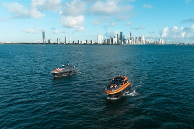 Two sleek motorboats cruising turquoise bay with Miami skyline and causeway bridge on the horizon under a sunny blue sky.