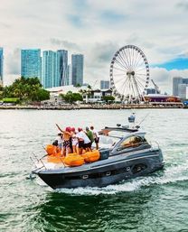 People celebrating on the bow of a sleek gray yacht with orange cushions cruising past Miami waterfront skyscrapers and a large Ferris wheel under a partly cloudy sky.