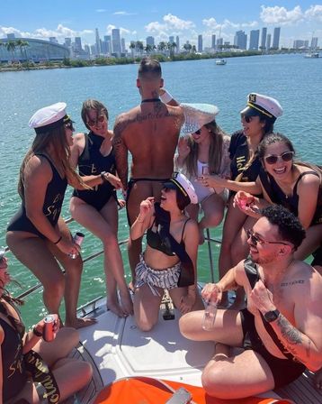 Laughing group of friends in swimsuits and captain hats on a sunny party boat near the Miami skyline, women with sashes and a man posing in a thong entertaining the crowd