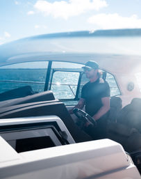 Man in cap and sunglasses cruising and steering a sleek motorboat on sunlit coastal waters, modern helm and cabin interior with ocean horizon visible.