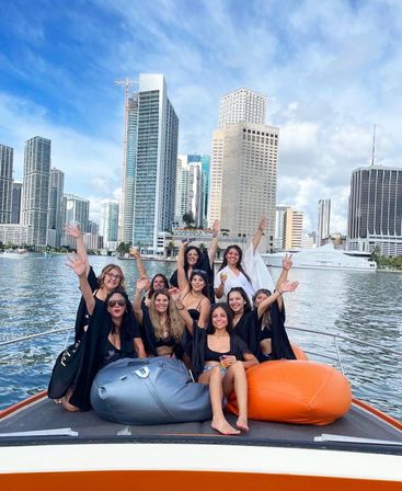 Group of women in swimsuits and black cover-ups cheering and waving from the bow of a boat with orange beanbags, set against downtown Miami high‑rise skyline and bright blue sky over the water.