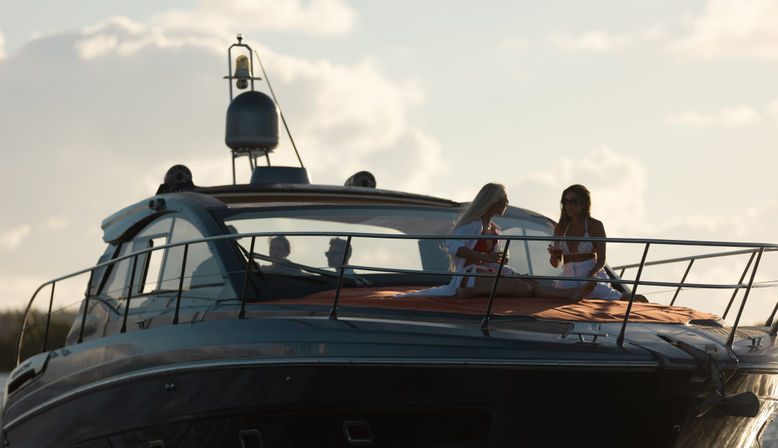 Two women in swimsuits enjoying drinks while lounging on the bow of a luxury motor yacht at sunset, silhouetted against a golden sky over open water.