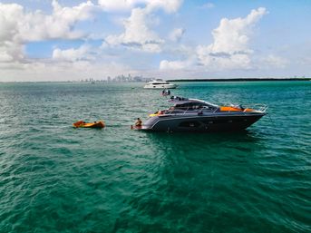 Luxury motor yacht anchored in turquoise coastal waters with a small orange kayak and person nearby, a second yacht and distant city skyline under a blue, cloudy sky.