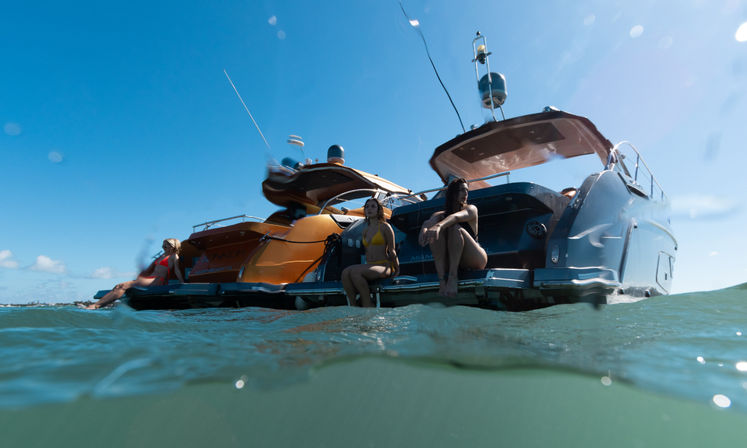 Two luxury yachts anchored in clear coastal water with three people in swimsuits relaxing on the swim platforms under a bright blue sky.