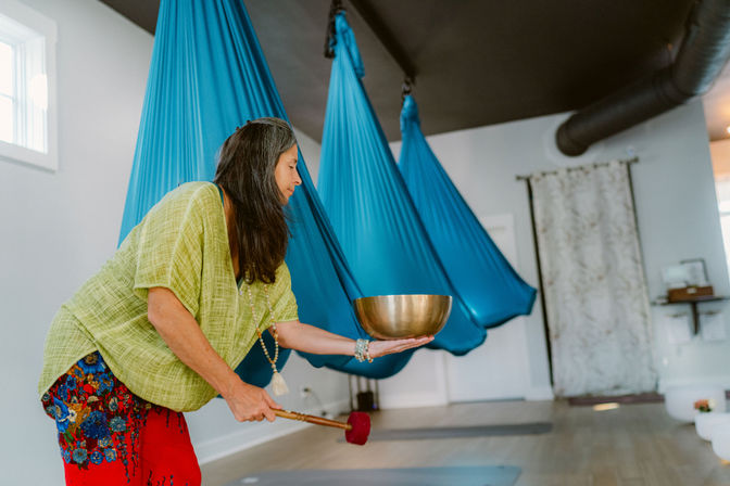 Aerial yoga studio interior with a person holding a brass singing bowl and mallet for sound healing, bright blue aerial hammocks and yoga mats on a wooden floor.