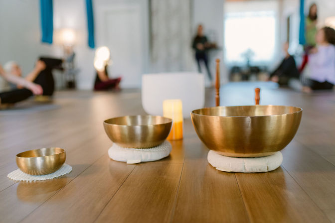 Three brass singing bowls on cushions with a candle on a wooden floor in a yoga studio sound bath meditation, blurred participants stretching in the background